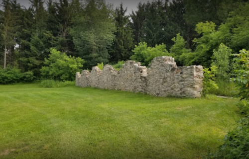 Stone ruins partially covered by greenery, set against a backdrop of trees and a grassy area.
