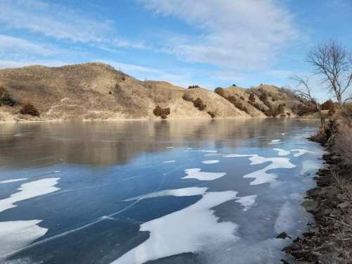 A serene winter landscape featuring a frozen lake, snow patches, and rolling hills under a blue sky.