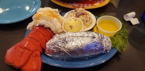 A plate featuring lobster tail, a baked potato wrapped in foil, coleslaw, butter, and a drizzle of olive oil on lettuce.