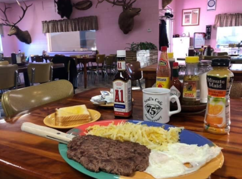 A plate with steak, eggs, and hash browns, surrounded by condiments and a cup, in a cozy diner setting.