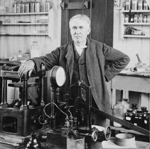 A historical black-and-white photo of a man in a lab, standing beside scientific equipment and instruments.