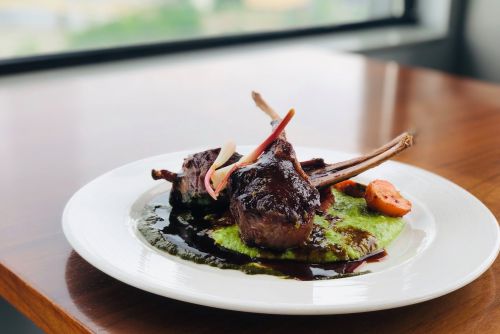 A plate of two lamb chops on a green puree, garnished with colorful vegetables and a rich sauce, set against a window view.