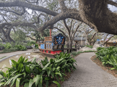 A colorful pirate ship playground surrounded by lush greenery and large trees in a park setting.