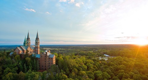 Aerial view of a church with tall spires, surrounded by lush greenery and a sunset in the background.