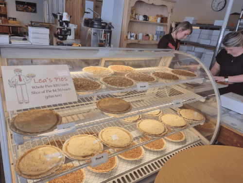 Display case of pies at Lea's Pies, featuring whole and sliced options, with prices listed. Two staff members in the background.