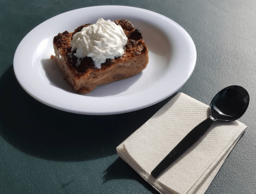 A slice of bread pudding topped with whipped cream, served on a white plate with a black spoon and napkin.