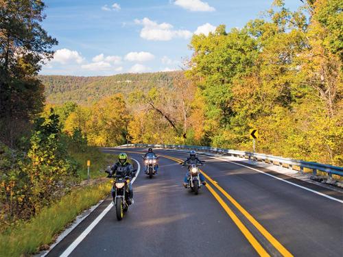 Three motorcycles ride along a winding road surrounded by vibrant autumn foliage and rolling hills.