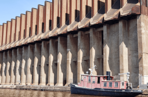 A small boat docked beside a large, industrial concrete structure with multiple columns and shadows.