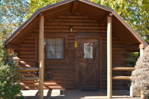 A rustic log cabin with a wooden door and porch, surrounded by greenery and trees.
