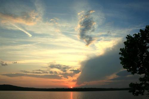 A serene sunset over a calm lake, with colorful clouds reflecting in the water and a silhouette of a tree in the foreground.