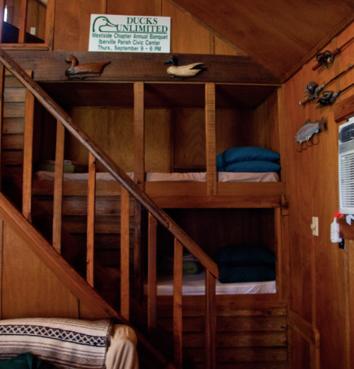 A wooden staircase leads to a cozy loft with stacked blankets and a sign for a Ducks Unlimited event on the wall.