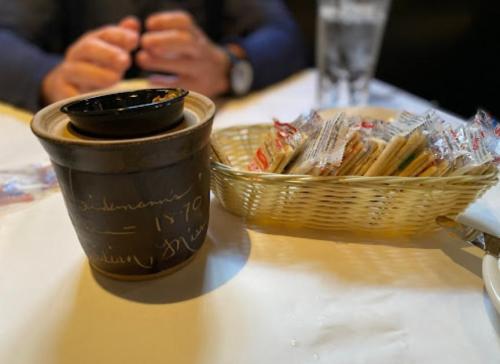 A clay pot with a lid sits on a table next to a basket of wrapped crackers and a glass of water.