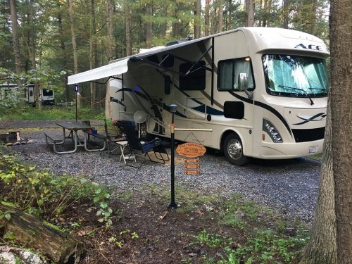 A parked RV in a wooded campsite, with a picnic table, chairs, and a sign nearby.