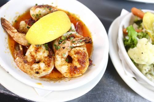 A bowl of shrimp in a spicy sauce, garnished with a lemon wedge, alongside a side of steamed vegetables.