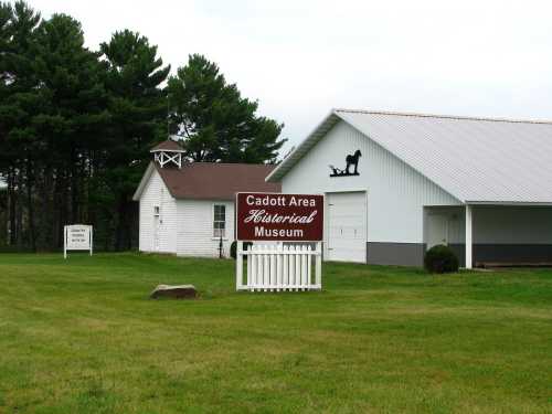 Sign for the Cadott Area Historical Museum in front of a white building, surrounded by green grass and trees.