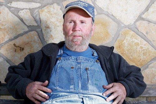 A man with a beard wearing a cap and overalls sits on a bench against a stone wall, looking thoughtfully ahead.