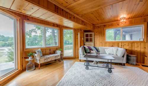 Cozy living room with wooden walls, large windows, a sofa, and a decorative rug, overlooking a green outdoor view.