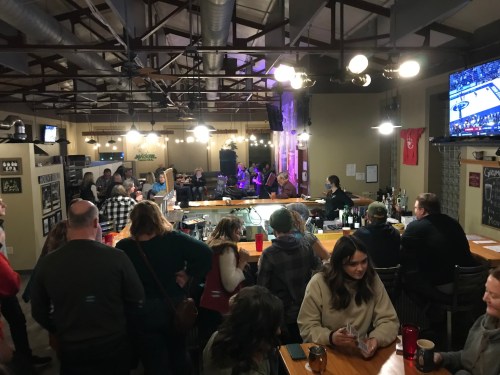 A lively bar scene with people socializing, a bar counter, and a sports game playing on a TV in the background.