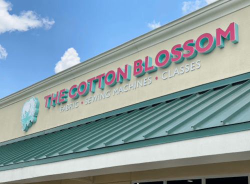 Sign for "The Cotton Blossom" store featuring fabric, sewing machines, and classes, with a blue sky in the background.