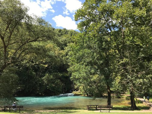 A serene river surrounded by lush green trees and a clear blue sky, with benches for visitors to enjoy the view.