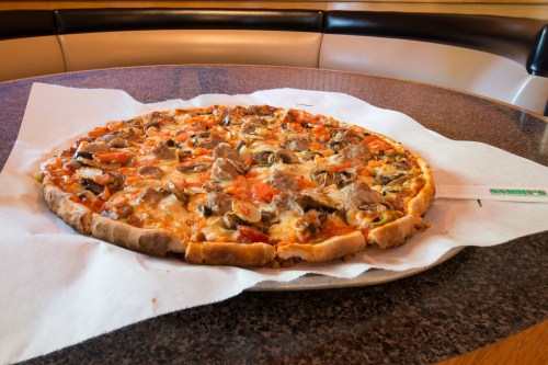 A large pizza topped with mushrooms, tomatoes, and meat, placed on a table with a white paper underneath.