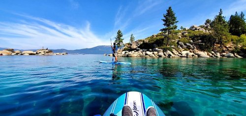 A person paddleboarding on clear blue water, surrounded by rocky shores and trees under a bright blue sky.