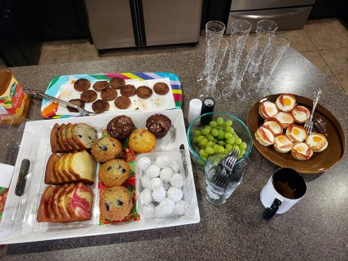 A table spread with various baked goods, fruits, and drinks, including muffins, cookies, and sliced cakes.