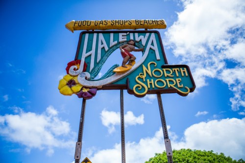 Colorful sign for Haleiwa, North Shore, featuring a surfer and flowers against a blue sky with clouds.