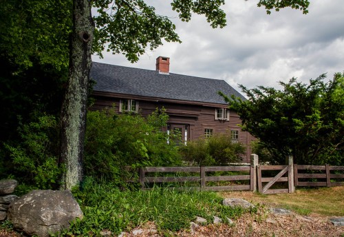 A rustic brown house surrounded by greenery and a wooden fence, under a cloudy sky.