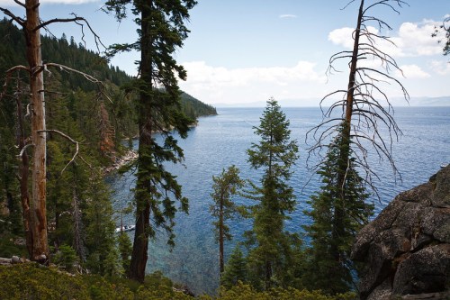 A serene view of a lake surrounded by tall trees and rocky shores under a partly cloudy sky.