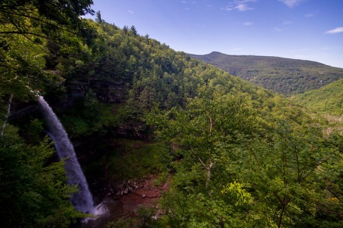 A scenic view of a waterfall cascading down a lush green hillside, surrounded by trees and mountains under a clear blue sky.