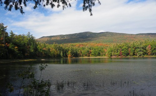 A serene lake surrounded by trees, with autumn foliage and mountains in the background under a partly cloudy sky.