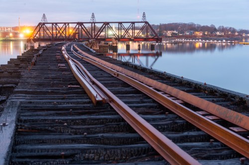 Rusty train tracks lead to a bridge over calm water, reflecting lights from nearby buildings at dusk.