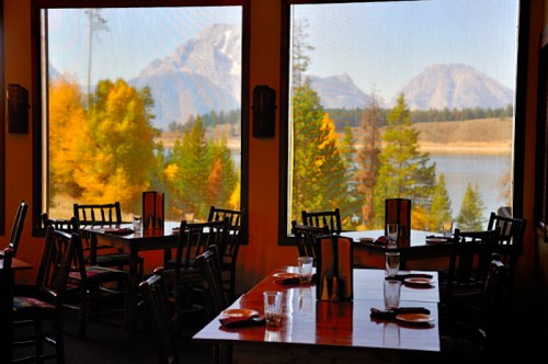 Cozy restaurant interior with wooden tables, overlooking vibrant autumn trees and mountains through large windows.