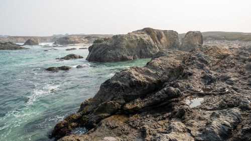 Rocky coastline with waves crashing against the shore, under a hazy sky.
