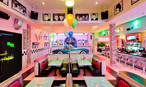 A vibrant retro diner with colorful balloons, booths, and a classic car on display, adorned with vintage photos.