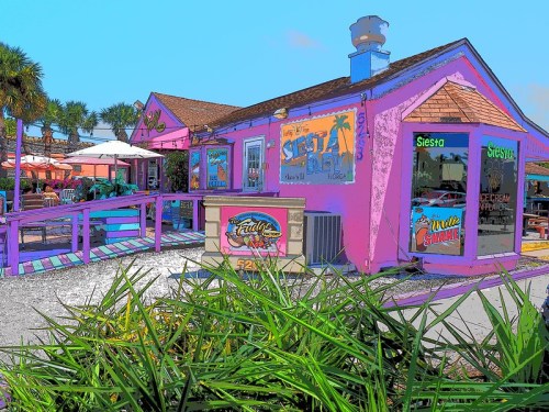 Colorful beachside restaurant with pink exterior, outdoor seating, and tropical decor surrounded by greenery.