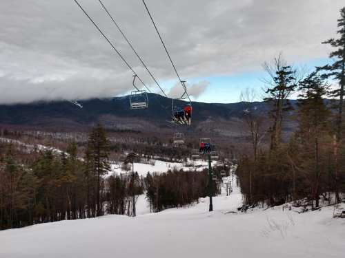 Two skiers on a chairlift above a snowy landscape with trees and mountains in the background under a cloudy sky.