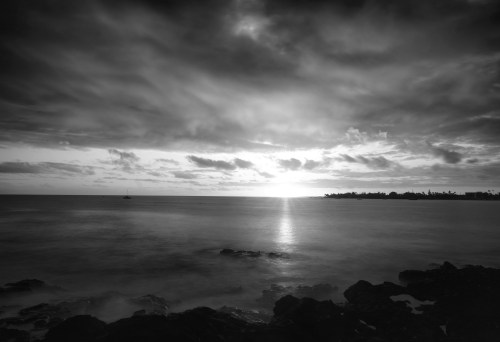 Black and white seascape at sunset, with clouds reflecting light over calm waters and rocky shoreline.