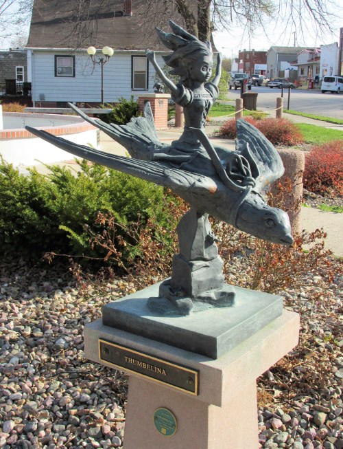 A bronze statue of Thumbelina riding a bird, mounted on a pedestal in a landscaped area.