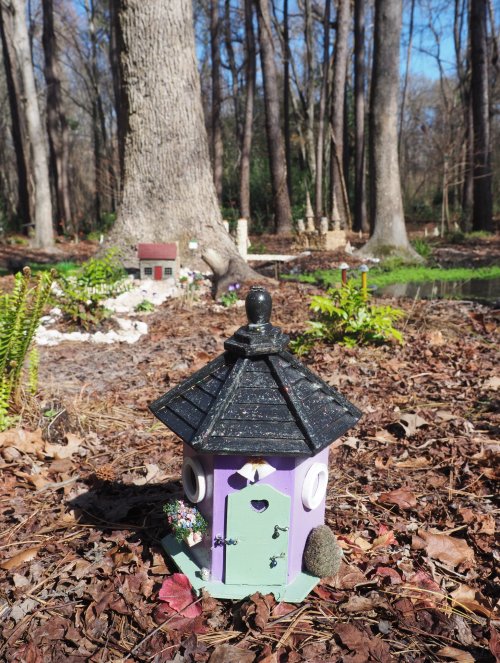 A whimsical purple birdhouse with a black roof, surrounded by fallen leaves and trees in a sunny garden setting.