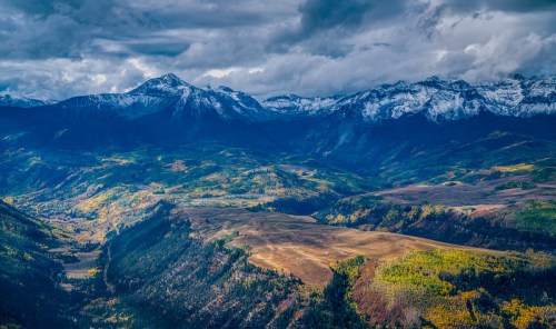 A panoramic view of snow-capped mountains and lush valleys under a dramatic, cloudy sky.