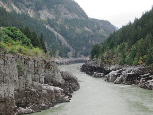 A winding river flows between steep, rocky cliffs, surrounded by lush green trees and mountains in the background.