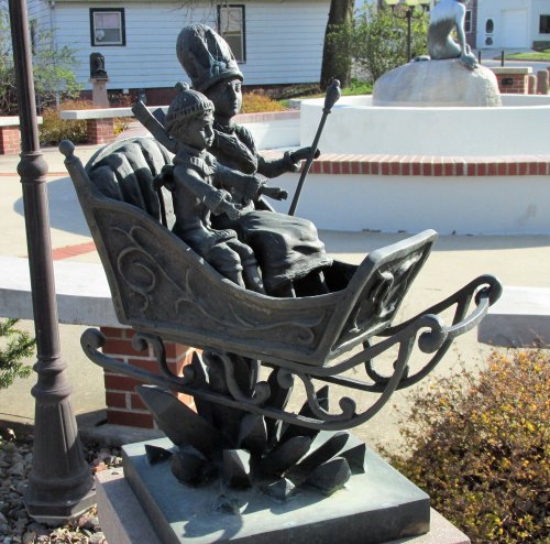 A bronze sculpture of a figure in a robe and a child, seated in a sleigh, surrounded by a park setting.