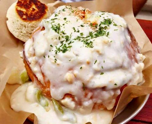 A large, creamy dish topped with herbs, served with a toasted bread roll on the side, all on a paper-lined plate.