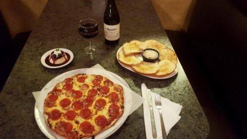 A table setting with a pepperoni pizza, garlic bread, a glass of red wine, and a dessert.