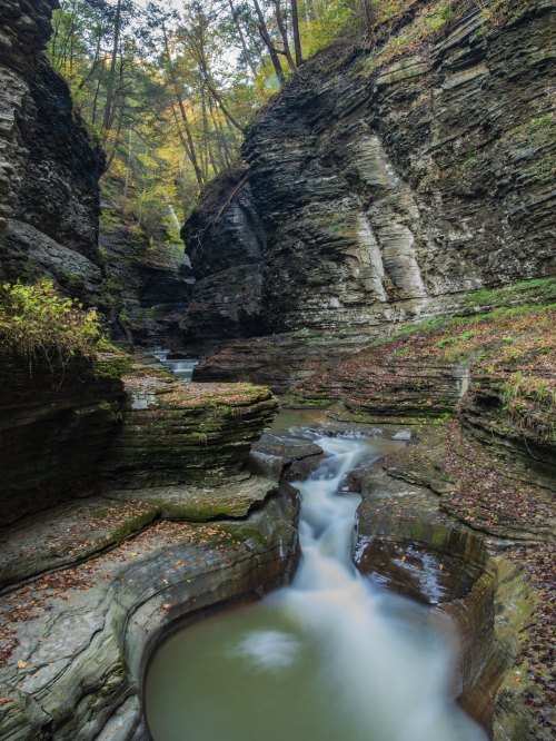 A serene river flows through a narrow canyon, surrounded by steep, rocky cliffs and autumn foliage.