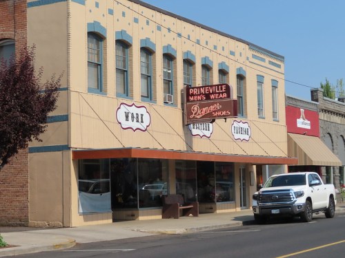 A storefront with "Prineville Men's Wear" and "Danner Shoes" signs, featuring large windows and a parked white truck.