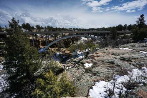 A scenic view of a bridge arching over a rocky landscape with patches of snow and trees under a cloudy sky.