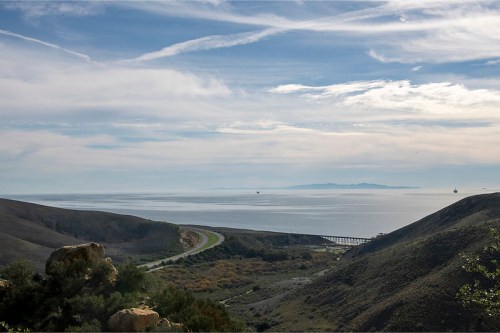 A scenic view of a coastline with rolling hills, a winding road, and a calm ocean under a cloudy sky.